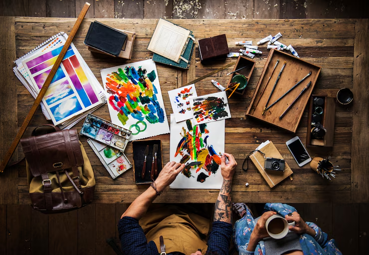 Overhead view of two people painting and drawing at a messy wooden table filled with art supplies, depicting creative work.