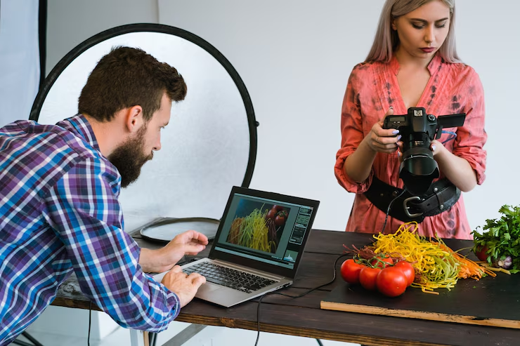 Two photographers setting up a food photoshoot with a camera, laptop, and colorful pasta, indicating culinary photography.