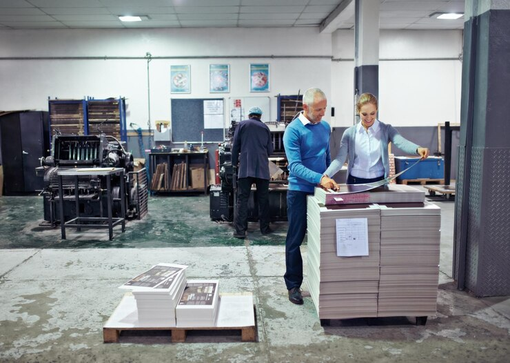 Two workers in a printing shop examining stacks of paper and machinery, showcasing industrial production or graphic design.